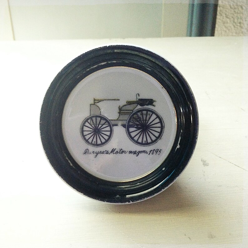 May include: A black and white image of a vintage car on a white porcelain plate with a black rim. The car is a Duryea's Motor Wagon, 1895.