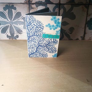 May include: A small, open booklet with a white cover featuring a blue coral design. The word "fear" is printed on a teal strip of paper. The booklet is on a wooden surface, with a patterned tile background.