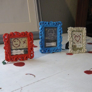 Może przedstawiać: Trzy małe obrazki w ramkach z kolorowymi obramowaniami. Ramki są czerwone, niebieskie i srebrne. Obrazki są wyszywane prostymi wzorami i tekstem. Tekst brzmi "Comedy", "Tragedy" i "Love".