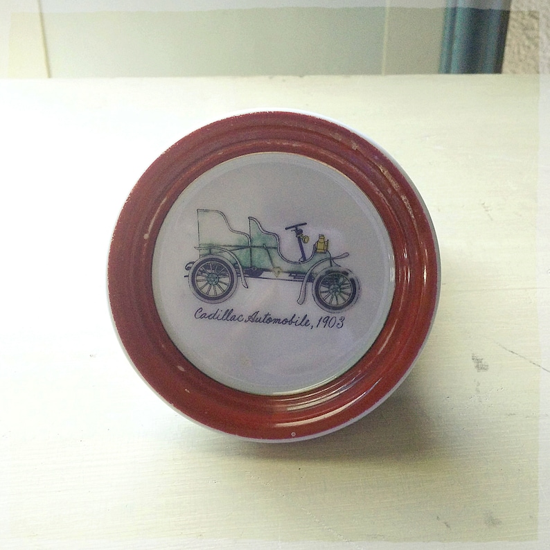 May include: A vintage porcelain plate with a red rim features a hand-painted illustration of a green 1903 Cadillac automobile. The text "Cadillac Automobile, 1903" is written below the car.