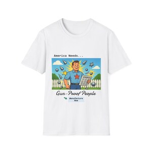 May include: White t-shirt with a graphic featuring a cartoon man holding a newspaper, surrounded by bullets and stars. Text reads "America Needs... Gun-Proof People" with "Manufacture Now" below.