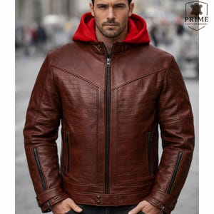 May include: A brown leather jacket featuring a textured crocodile-embossed pattern on the chest and sleeves, a full-length black front zipper, and a contrasting bright red hood. The jacket includes zippered side pockets and adjustable cuffs.