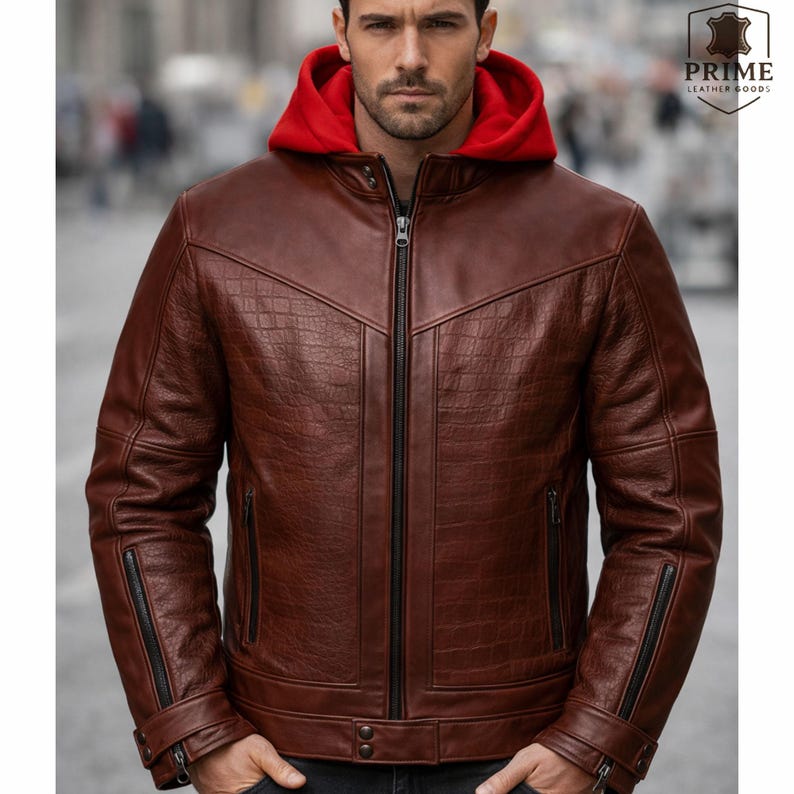May include: A brown leather jacket featuring a textured crocodile-embossed pattern on the chest and sleeves, a full-length black front zipper, and a contrasting bright red hood. The jacket includes zippered side pockets and adjustable cuffs.
