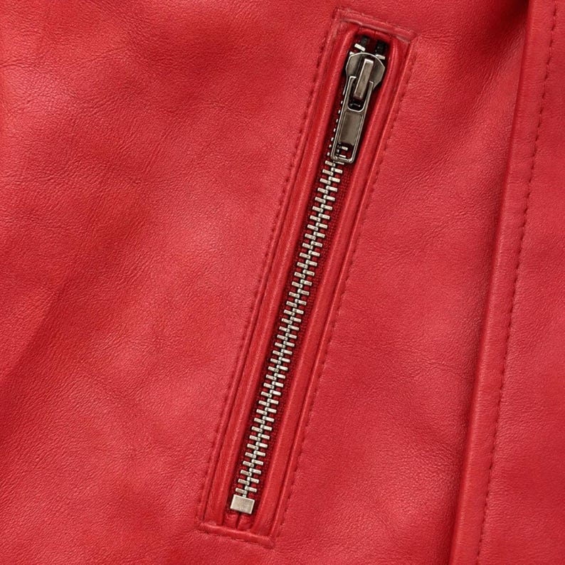 May include: A bright red jacket featuring a smooth, textured surface and a vertical zippered pocket. The pocket is secured with a metallic silver zipper pull and teeth, set against the vibrant red material with visible stitched seam detailing.