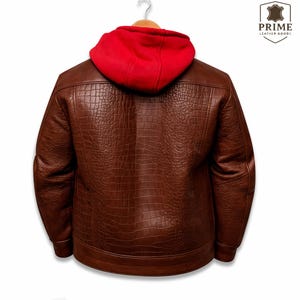 May include: A brown leather jacket with a textured crocodile skin pattern and a bright red fabric hood. The jacket is displayed on a hanger and features the Prime Leather Goods logo in the upper right corner with a shield icon and text.