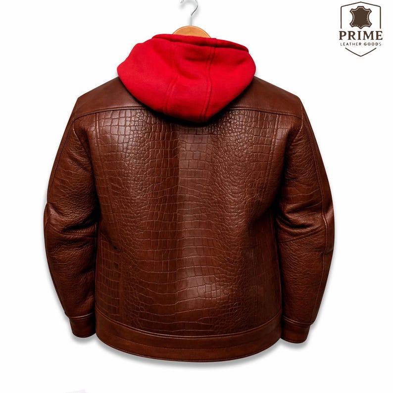 May include: A brown leather jacket with a textured crocodile skin pattern and a bright red fabric hood. The jacket is displayed on a hanger and features the Prime Leather Goods logo in the upper right corner with a shield icon and text.