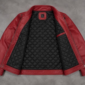 May include: A bright red jacket with a stand-up collar and long sleeves. The interior features a black quilted lining with a diamond pattern. A rectangular label with the letter L is centered at the inner neckline. The jacket has metal zippers.