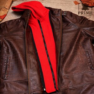 May include: A brown leather jacket with a textured crocodile-embossed pattern, featuring a detachable bright red hooded inner layer with a black zipper closure. The jacket includes snap button details on the collar and a logo reading PRIME LEATHER GOODS.