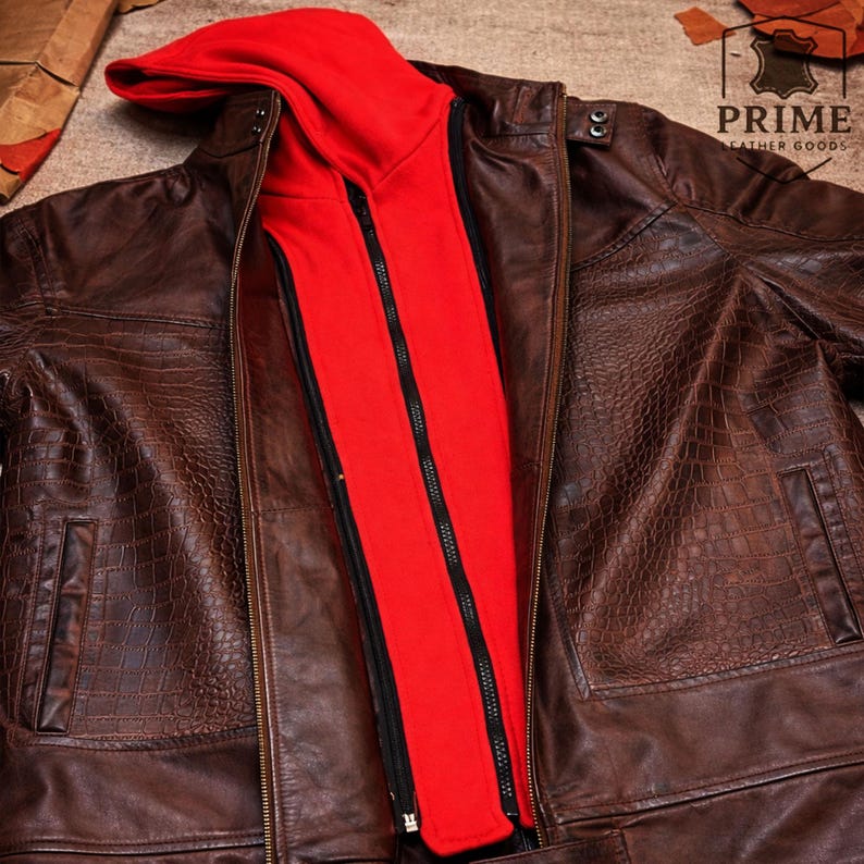 May include: A brown leather jacket with a textured crocodile-embossed pattern, featuring a detachable bright red hooded inner layer with a black zipper closure. The jacket includes snap button details on the collar and a logo reading PRIME LEATHER GOODS.