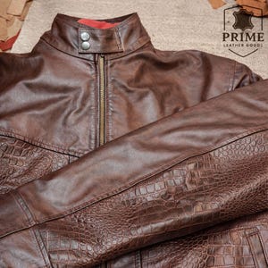May include: A brown leather jacket featuring a stand-up collar with two silver-tone snap buttons, a central brass-colored zipper, and a textured crocodile-embossed pattern on the lower sections. The logo PRIME LEATHER GOODS is visible.