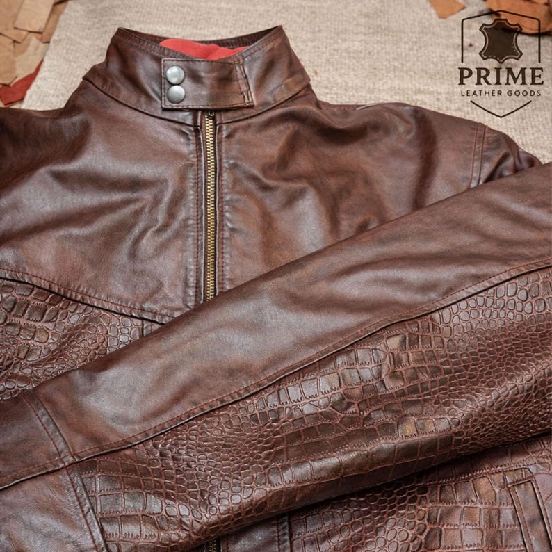 May include: A brown leather jacket featuring a stand-up collar with two silver-tone snap buttons, a central brass-colored zipper, and a textured crocodile-embossed pattern on the lower sections. The logo PRIME LEATHER GOODS is visible.