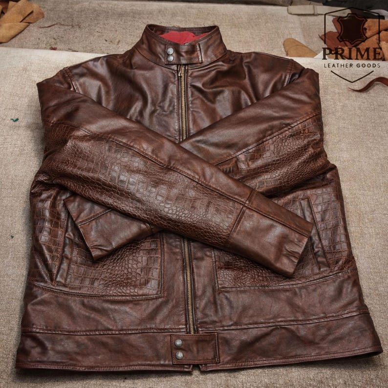 May include: A brown leather jacket featuring a stand-up collar with two snap buttons, a full-length front zipper, and textured crocodile-embossed panels on the sleeves and lower front pockets. The interior lining is a vibrant red fabric.