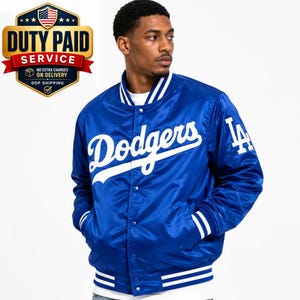 Op de afbeelding: Een koningsblauw satijnen varsity-jack met het witte Dodgers-schriftlogo op de borst en het LA-logo op de mouw. Het jack heeft een drukknoopsluiting aan de voorzijde, een gestreepte ribgebreide kraag en gestreepte ribgebreide boorden in wit en blauw.