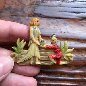 May include: A vintage brooch featuring a couple in a romantic scene. The woman is standing with a bouquet of flowers, while the man is kneeling down on one knee. The brooch is made of white and cream colored plastic with painted details in red, green, and blue.