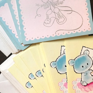 May include: A stack of blue and yellow baby shower cards with envelopes. The cards feature a blue teddy bear holding a sign that says "A New Baby".