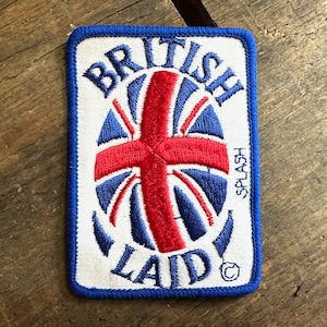 May include: A white patch with blue edging and a red, white, and blue embroidered Union Jack design. The text "BRITISH LAID" is embroidered around the Union Jack design. The text "SPLASH" is embroidered vertically on the right side of the patch.