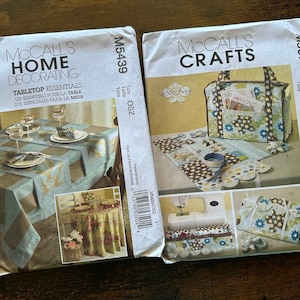 May include: Two sewing patterns for home decor projects. The first pattern, McCall's Home Decorating M5439, shows a table setting with a blue and brown tablecloth and napkins. The second pattern, McCall's Crafts M5871, shows a tote bag, a table runner, and a small pouch, all with floral fabric.