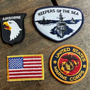 May include: Four embroidered patches featuring military themes. The patches are black, blue, red, and yellow. The patches feature an eagle, a ship, an American flag, and the United States Marine Corps emblem.
