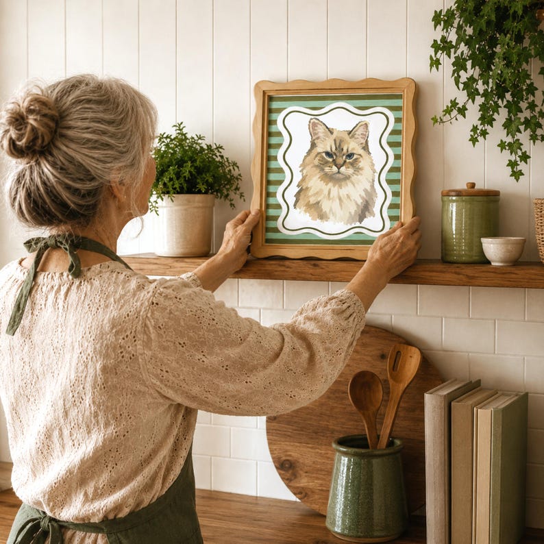 May include: A square wooden picture frame with a scalloped edge containing a print of a fluffy, light brown cat. The background of the print features horizontal green and white stripes, and the cat is centered within a white, wavy-edged border.