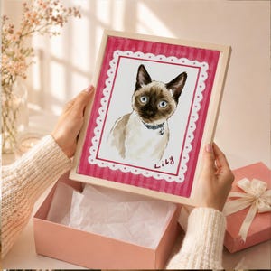 May include: A framed watercolor illustration of a Siamese cat with blue eyes and a dark face mask, wearing a collar. The artwork features a white scalloped border on a pink striped background with the handwritten name Lily in the bottom corner.