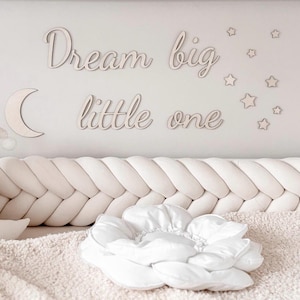 Könnte beinhalten: Eine Szene im Kinderzimmer mit einem cremefarbenen geflochtenen Stoßfänger, einem Mond aus Plüsch und einem weißen blütenförmigen Kissen. An der Wand befinden sich Holzausschnitte von Sternen, einem Mond und dem Text "Dream big little one."