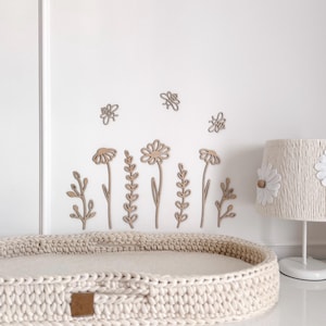 Peut inclure: Une scène de chambre d'enfant avec un panier à langer en crochet beige, une lampe blanche avec des détails de marguerites et des décorations murales florales en bois. L'art mural comprend des fleurs et des abeilles. La scène est contre un mur blanc et une commode blanche.