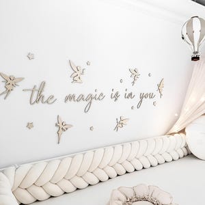 Può includere: Una scena di decorazione murale per la cameretta con ritagli di legno a forma di fata e stella, e la frase "the magic is in you". Sono visibili anche un paracolpi intrecciato e un cuscino a forma di fiore. Una mongolfiera decorativa è appesa al soffitto.