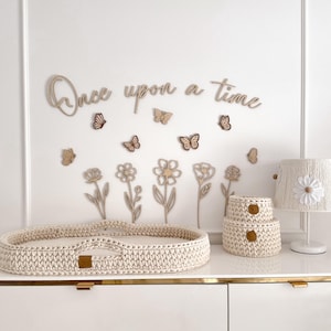 Può includere: Una scena di cameretta con un comò bianco, con un cestino fasciatoio intrecciato, due scatole portaoggetti abbinate e una lampada. La parete è decorata con fiori di legno, farfalle e la frase "Once upon a time."