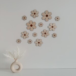 May include: A collection of wooden flower cutouts in various sizes, each with a light brown petal and a darker brown center. The flowers are arranged on a white wall, creating a decorative floral pattern. A vase with white decorative grass is in the foreground.
