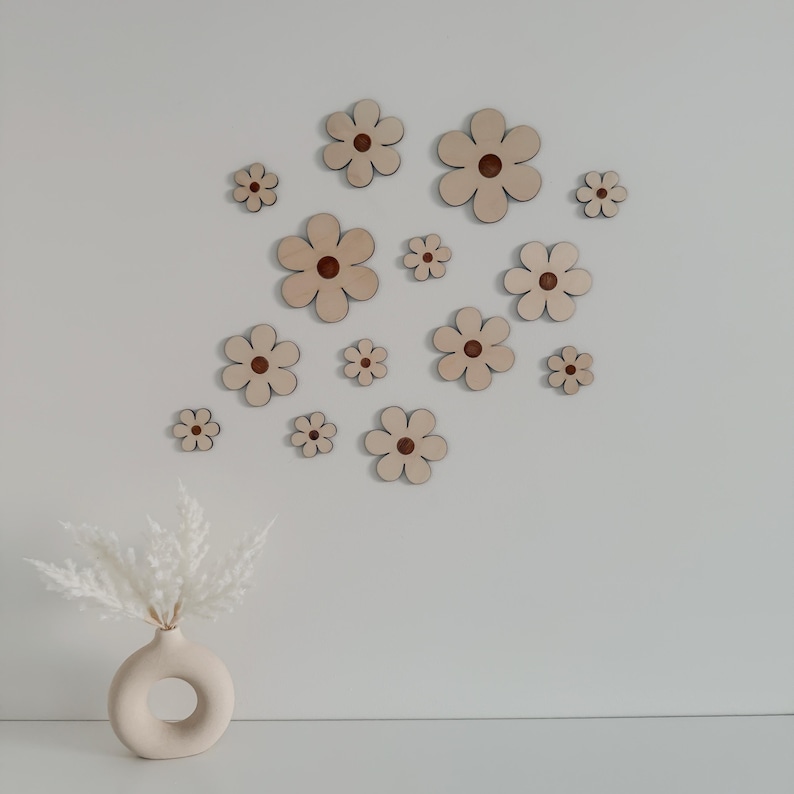 May include: A collection of wooden flower cutouts in various sizes, each with a light brown petal and a darker brown center. The flowers are arranged on a white wall, creating a decorative floral pattern. A vase with white decorative grass is in the foreground.
