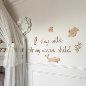 Könnte beinhalten: Wanddekoration für das Kinderzimmer mit Holzausschnitten von Meeresbewohnern und dem Schriftzug "stay wild my ocean child". Das Design umfasst ein Seepferdchen, einen Seestern, eine Muschel, eine Schildkröte, einen Wal und Korallen.