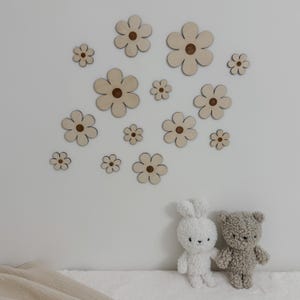 May include: Wooden flower wall decor in various sizes, with a light wood finish and a darker center. The flowers are arranged on a white wall. A white bunny and a gray teddy bear plush toy are in the foreground.