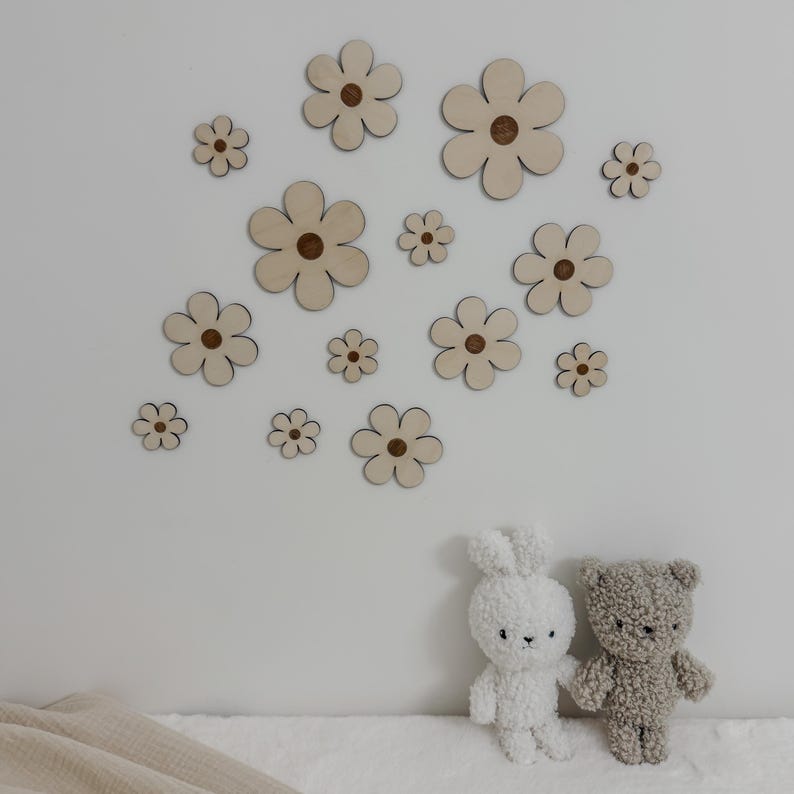 May include: Wooden flower wall decor in various sizes, with a light wood finish and a darker center. The flowers are arranged on a white wall. A white bunny and a gray teddy bear plush toy are in the foreground.