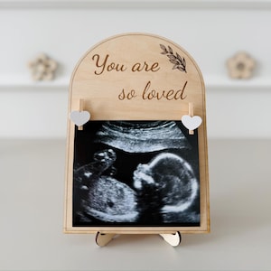 May include: Wooden pregnancy announcement frame with the text "You are so loved" and a sonogram image. The frame has a decorative arch top and is supported by a small stand. Two heart-shaped clips hold the sonogram in place.