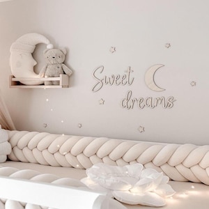 Op de afbeelding: Een neutrale kinderkamer met een crèmekleurige gevlochten bumper, een witte wieg en wanddecoratie met een maanvormig rek met een teddybeer en een halve maan kussen. De muur heeft ook de woorden "Sweet dreams" en steraccenten.