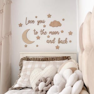 Peut inclure: Décoration murale de chambre d'enfant avec des découpes en bois d'un croissant de lune, d'étoiles et d'un nuage, avec l'inscription « Love you to the moon and back ». Le lit est orné d'un oreiller à carreaux et de coussins en peluche décoratifs.