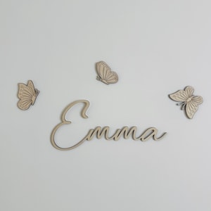 Könnte beinhalten: Der Name "Emma" aus Holz in Schreibschrift, zusammen mit drei Holz-Schmetterlingsdekorationen. Die Schmetterlinge und der Name sind hellbraun vor weißem Hintergrund und bilden eine einfache, dekorative Anordnung.