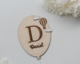 Może przedstawiać: Drewniana dekoracja w kształcie balonu z literą "D" i wygrawerowanym imieniem "Daniel". Projekt zawiera balon na ogrzane powietrze i chmury. Biały sznurek jest przymocowany na dole.