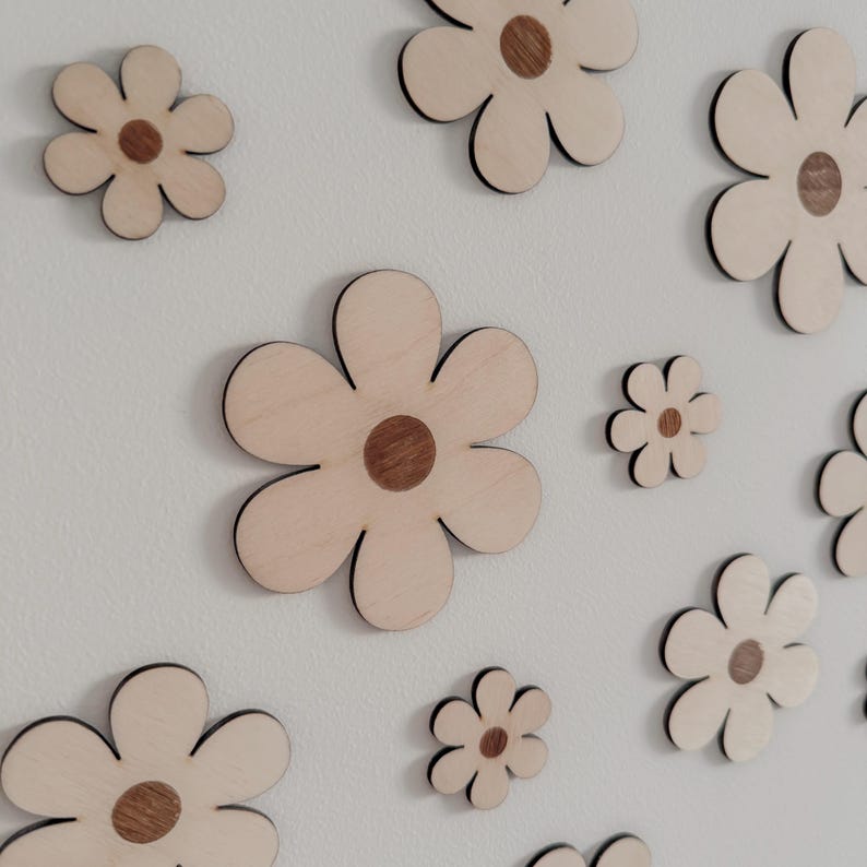 May include: Wooden flower cutouts in various sizes are arranged on a white surface. Each flower has a light wood petal design with a darker wood center. The flowers are a decorative element.