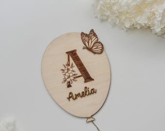 Może przedstawiać: Drewniana dekoracja w kształcie balonu z wygrawerowaną literą "A" i imieniem "Amelia". W literę wkomponowano kwiatowy wzór, a do niej przymocowano drewnianego motyla. Do dołu przymocowany jest sznurek.