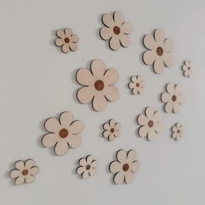 May include: A collection of wooden flower cutouts in various sizes, affixed to a white wall. Each flower features a light beige petal design with a dark brown center. The arrangement creates a whimsical, decorative wall art piece.
