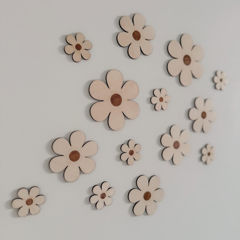 May include: A collection of wooden flower cutouts in various sizes, affixed to a white wall. Each flower features a light beige petal design with a dark brown center. The arrangement creates a whimsical, decorative wall art piece.