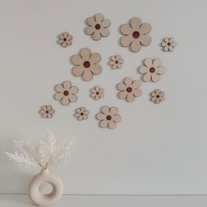May include: A collection of wooden flower wall decor in various sizes, arranged on a white wall. Each flower has a light wood tone with a dark brown center. A beige vase with white pampas grass is in the foreground.