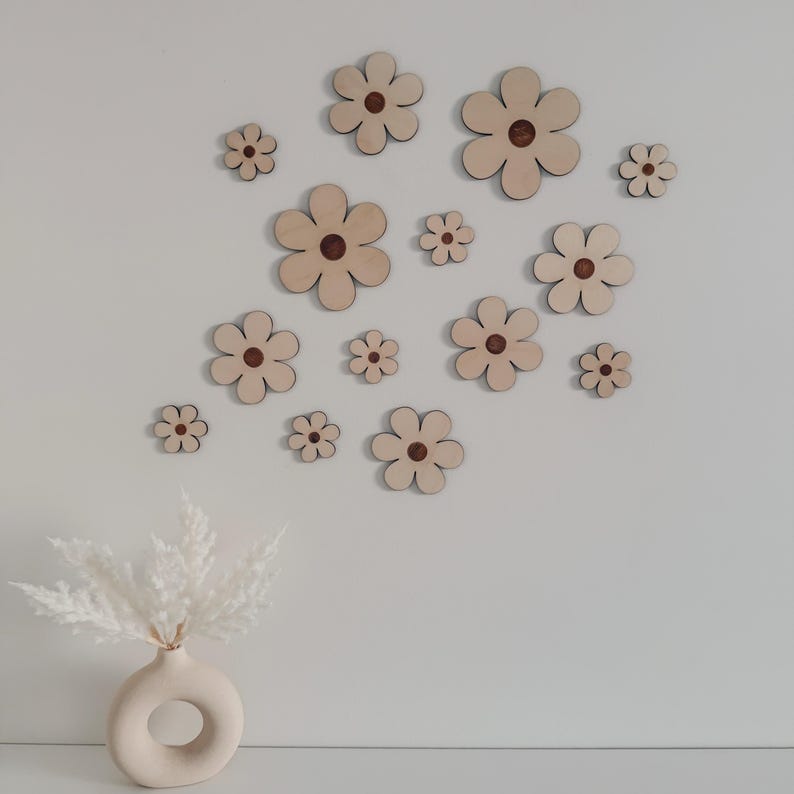 May include: A collection of wooden flower wall decor in various sizes, arranged on a white wall. Each flower has a light wood tone with a dark brown center. A beige vase with white pampas grass is in the foreground.