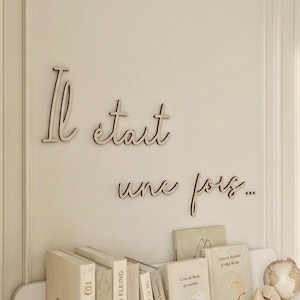 May include: Light-colored wooden wall lettering in a cursive script that reads "Il était une fois..." mounted above a shelf containing several books with neutral-toned covers, small wooden geometric blocks, and a miniature globe.
