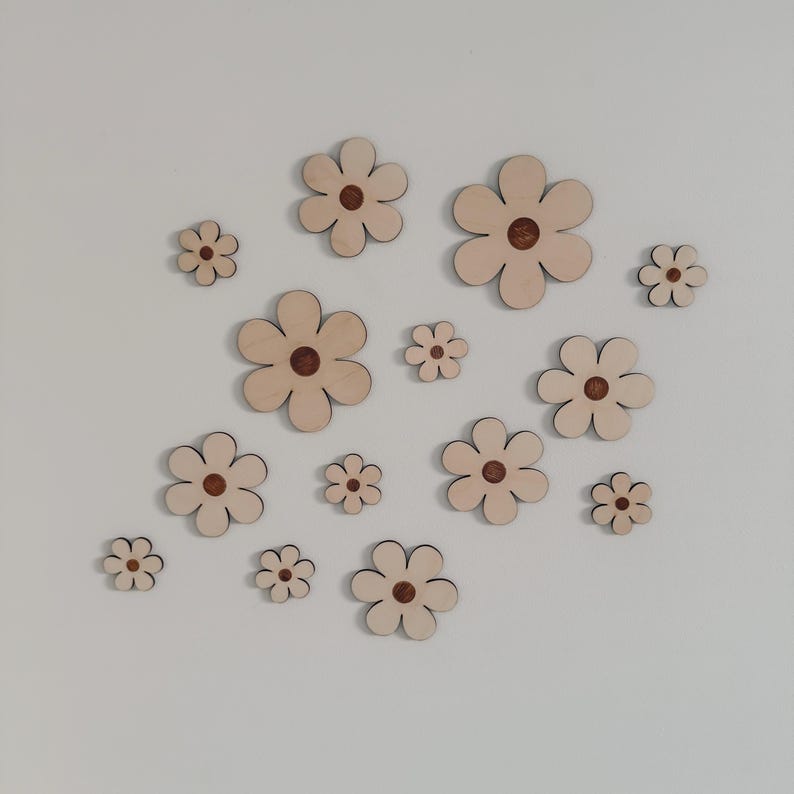 May include: A collection of wooden flower cutouts in various sizes. The flowers have a light beige color with a dark brown center. The cutouts are arranged on a white background, creating a decorative floral pattern.
