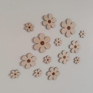 May include: A collection of wooden flower cutouts in various sizes, arranged on a white wall. The flowers have a light wood color with a darker brown center and a black outline, creating a simple, yet elegant design. These decorative flowers are perfect for wall decor.