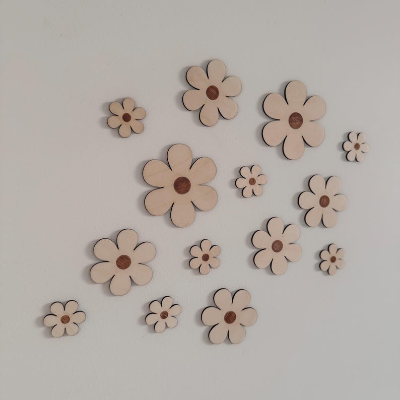 May include: A collection of wooden flower cutouts in various sizes, arranged on a white wall. The flowers have a light wood color with a darker brown center and a black outline, creating a simple, yet elegant design. These decorative flowers are perfect for wall decor.