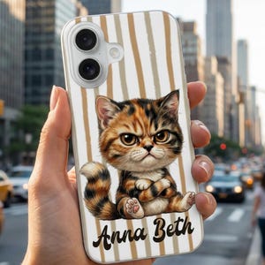 May include: A clear phone case featuring a cartoon cat with orange, brown, and black fur, and the name "Anna Beth" in a script font. The background has vertical beige and white stripes.