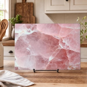 Op de afbeelding: Een rechthoekige glazen snijplank met een hoge resolutie print van roze rozenkwarts met witte aders. De plank wordt ondersteund door een zwarte metalen standaard en heeft een glad, gepolijst oppervlak met een kristallijne textuur.